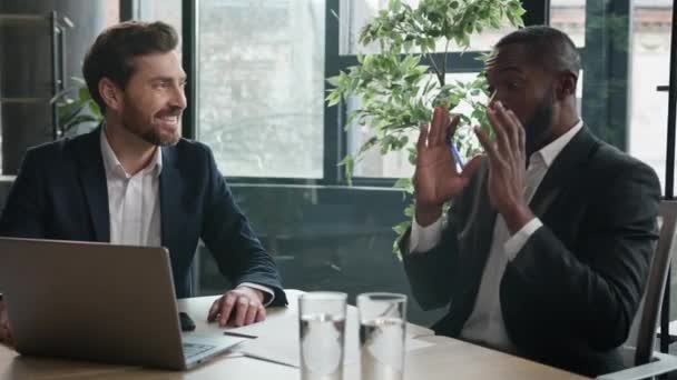 アフリカ人とコーカサス人のビジネスマンとのオフィスミーティングで笑って話す2人の多様なビジネスマンは 仕事でラップトップのパートナーシップの友人との友人との友好的なコワーキングで冗談を言います — ストック動画