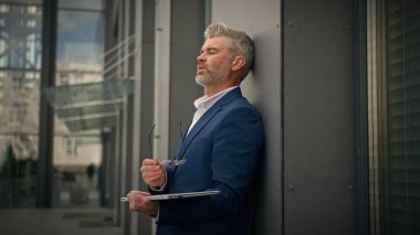 Yorgun, Kafkasyalı, yaşlı işadamı, olgun iş adamı, dizüstü bilgisayarı olan orta yaşlı, yönetici, iş bitirici şehir dışında, ofis binasının duvarına yaslanmış gözlükleri çıkarıyor.