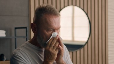 Ev banyosunda hasta olan 60 'lı yaşlardaki yaşlı beyaz bir adam kağıt peçeteyle burnunu silerken yaşlı, yaşlı, hasta, yaşlı bir erkek dedesi burun nezlesi belirtileri göstererek hapşırıyor.