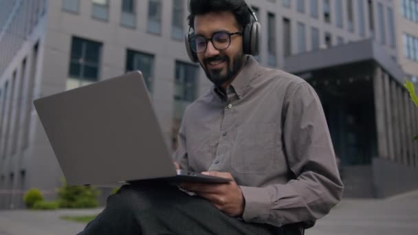アラビアのインディアン男性は ヘッドフォンで音楽教育ウェビナーを聞きます 男性のビジネスマンフリーランサーは ノートパソコンでビジネスプロジェクトを開発する 都市で笑顔の男の学生Eラーニングをオンラインで勉強 — ストック動画