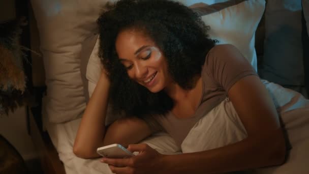 非洲裔美国人快乐女人沉溺于床上手机滚动网络社交媒体智能手机小工具成瘾的少数民族女孩在卧室浏览网上约会应用程序 — 图库视频影像