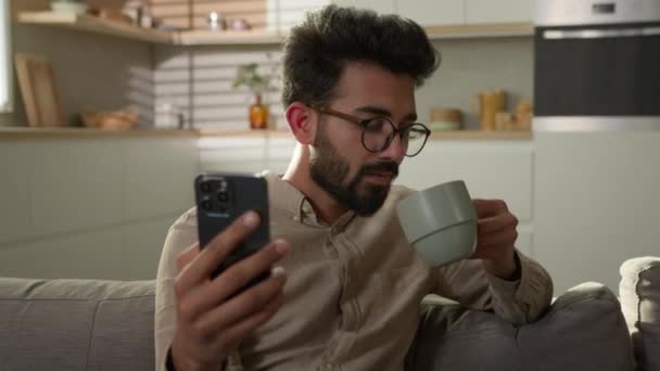 在家里厨房里 男性快乐快乐的家伙在网上看电视片 在手机上看新闻 喝早茶 在沙发上放松地看着相机微笑 — 图库视频影像
