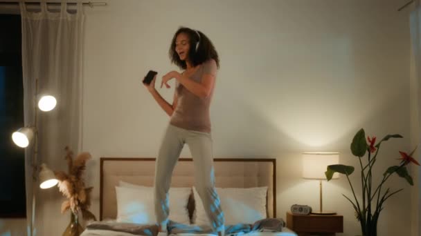 无忧无虑地嬉闹着的非洲裔美国女孩在床上跳着玩 欢度假期 欢快地跳舞 在家里聚会 用手机唱歌 用耳机听音乐 — 图库视频影像