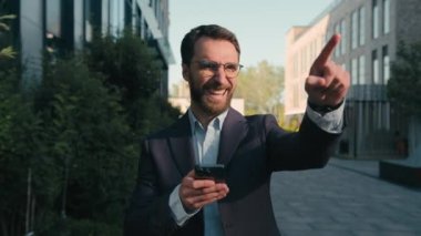 Gülümseyen mutlu beyaz erkek işadamı şehir caddesinde geziyor mobil uygulamalarla sohbet ediyor heyecanlı erkek müşteriler ofis binasını işaret ediyor telefon akıllı telefonunu açık havada kullanıyor.