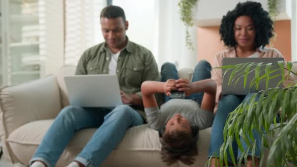 吸毒成瘾者家庭使用家用电器小男孩倒地躺在沙发上玩电子游戏手机微笑着非裔美国父母使用现代技术在不同的笔记本电脑上工作 — 图库视频影像