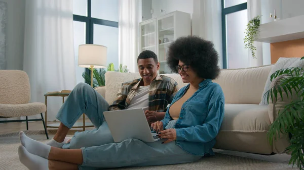 African American couple on floor at home using online laptop computer browsing shopping together pay bills choose products order delivery do high five gesture teamwork happy family in own apartment