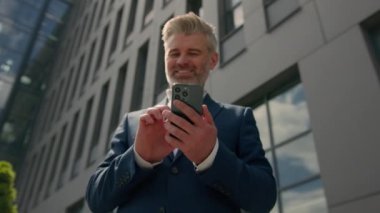 Kafkasyalı yetişkin 60 'lı yaşlarda mutlu bir işadamı mutlu bir işadamı şehrin dışında modern akıllı telefon üzerinde çalışıyor cep telefonlu olgun orta yaşlı iş adamına bak online bankacılık uygulaması e-işi ile sohbet ediyor