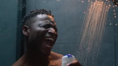 Afro-Amerikalı adam banyoda şarkı söylüyor, neşeli, mutlu, kaslı, atletik erkek enerjisi müzik eşliğinde dans ediyor şampuan banyosu jeliyle şarkı söylüyor, vücut bakımını tazeliyor.