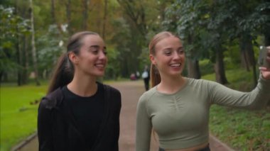 İki mutlu Kafkasyalı kız blogu kadınları cep telefonlu selfie kameralı cep telefonuyla video blogu kaydediyor. Şehir parkında yürüyüş yapıyor ve akıllı telefonları arıyor. Kadın sporcular dışarıda online fitness sporu yapıyor.