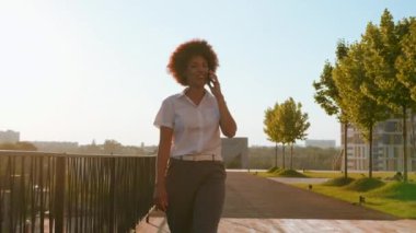 Mutlu gülümseyen Afro-Amerikan kıvırcık kadın konuşan cep telefonu gülümseyen açık havada yürüyen iş kızı internetten akıllı telefon konuşması yapan sohbet eden iş kadını şehir güneşinde yürüyen iş kadını.