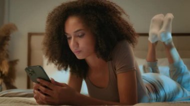 Afro-Amerikalı kadın etnik kız, genç kız, cep telefonu ağlarını kaydırıyor, gece yatak odasında uzanıyor, sohbet ediyor, akıllı telefon aygıtı aldatmacası hakkında kötü haberler okuyor.