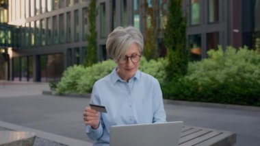 Yetişkin Kafkasyalı iş kadını banka kartı ve dizüstü bilgisayarla faturaları ödüyor. Yaşlı kadın yatırımcı, şehirde internet üzerinden para transferi bankası satın alıyor.