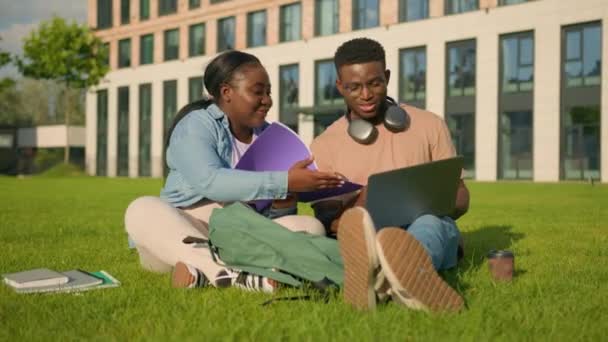 公園の2人のアフリカ系アメリカ人の友達の高校生が クラスメイトと一緒に勉強する大学の大学の外で 芝生の女の子のラップトップで学ぶことは 笑う宿題を持つ男を助けることを説明します — ストック動画