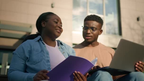 两名非洲裔美国朋友高中学生户外大学大楼与同学一起学习用笔记本电脑讨论网上作业女生用试卷解释帮助男生笑 — 图库视频影像