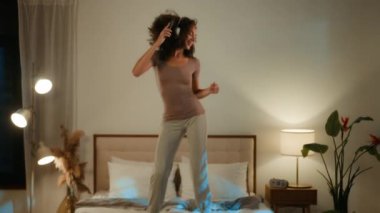 Genç, mutlu, Afro-Amerikalı, aktif, enerjik bir genç kız gece yatak odasında lamba ışığında parti eğlencesi eşliğinde cep telefonuyla dans eden kulaklıkla müzik dinleyerek yatağa atlıyor.