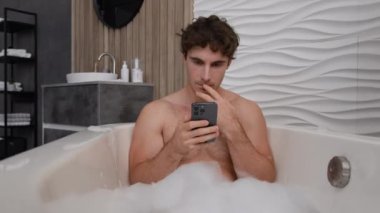 Kafkasyalı, sakin, sakin, çıplak, rahat, erkek, banyo küvetinde sabunlu, sıcak su banyosu yapan, akıllı telefon kullanan, sosyal medya uygulaması okuyan, düşünceli, düşünceli bir adam.