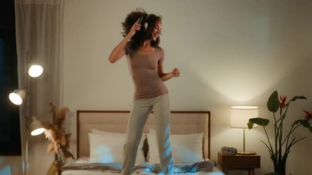 ジャンプ幸せな少女アフリカ系アメリカ人若い女性アクティブエネルギッシュな女性ティーンは ランプライトホームパーティーエンターテイメント携帯電話ダンスでヘッドフォンで音楽を聞く夜寝室でベッドにジャンプ — ストック動画