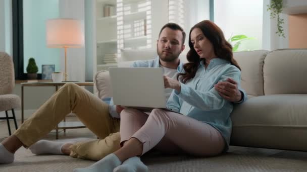 ハッピーファミリーコーカサス若い夫婦 リビングルームのホームフロアで夫妻 遠くのコンピュータを購入するコンピュータを見て一緒にオンラインショッピングでラップトップのEコマースを使用して選択することを議論 — ストック動画