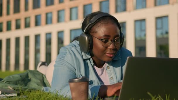 ハッピー アフリカ系アメリカ人女性 芝生に横たわる ワイヤレス コンピューター リスニング ヘッドフォン 研究ノートパソコン 屋外で遠くに勉強 テイクアウト — ストック動画
