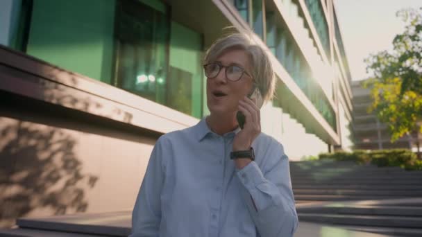 快乐的白种人成熟的老女商人女经理办公室女经理讲手机电话洽谈在城市里谈笑风生60多岁的女人聊天智能手机在户外晃来晃去 — 图库视频影像