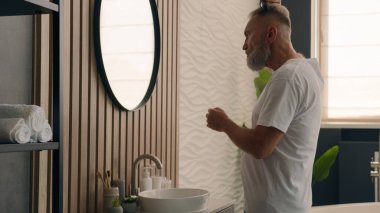 Banyodaki yakışıklı, kendine güvenen beyaz adam aynadaki yansımasına bakıyor. Sakal tarıyor, saçlarını tarıyor. Yaşlı bir erkek, saçlarını tarıyor.