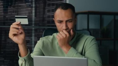 Kafası karışmış Afrikalı Amerikalı adam internetteki ödeme bankası iflası yüzünden bilgisayardan uzak kredi kartı kullanıyor. Sinirli erkek, yanlış şifre girerek elektronik hesaba aşırı para harcamaktan endişeleniyor.