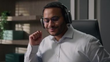 Mutlu Arap Hindistanlı çok ırklı Amerikalı iş adamı iş yerinde kulaklıkla müzik dinliyor dizüstü bilgisayarla dans eden iş adamı bilgisayarda dans edip şarkı söylüyor.