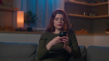 Gülümseyen mutlu beyaz kadın akşam kanepesinde boş vakitlerini cep telefonuyla sosyal ağda gezinerek e-posta alışverişini kontrol ederek geçiriyor akıllı telefondaki online mağazada alışveriş yapıyor.