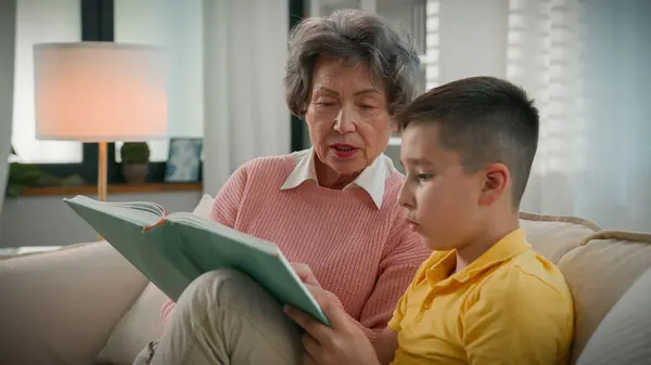 Loving elderly woman retired grandmother reading book with grandson at home on sofa enjoying read interesting fairytale story caring grandma helping child kid boy with homework studying encyclopedia