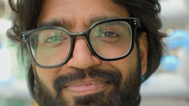 Close up positive male face in eyeglasses. Portrait Indian bearded man in glasses smiling looking at camera. Headshot entrepreneur guy businessman executive indoor with lights reflection in spectacles