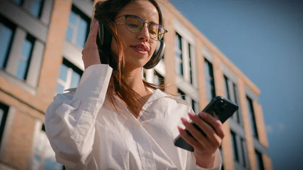 Happy woman in headphones listening audio music on phone outdoors in sunlight near office building carefree girl female using mobile application for enjoy favorite song dancing resting on urban street