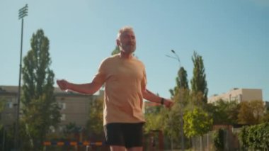 Beyaz, yaşlı bir adam, şehir stadyumunda ip atlarken fiziksel aktivite atlarken ısınma egzersizleri yapıyor. Eski sporcu, dışarıda vücut sağlığı için spor yapıyor.