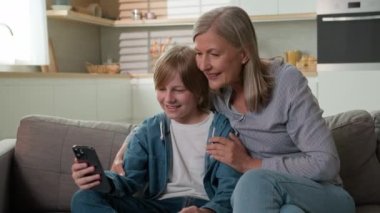 Sevgili büyükannemin torunuyla evde vakit geçirmesi olgun kadın büyükanne ile küçük çocuk arasında internette komik videolar izlemesi iki nesil boyunca akıllı telefon kullanarak mutlu Kafkas ailesi.