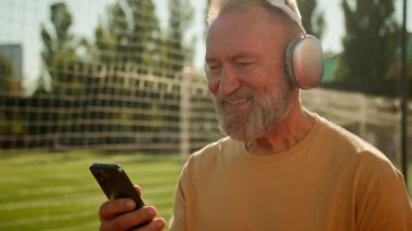 Beyaz, mutlu, yaşlı bir adam stadyum futbol sahasında kulaklıklarla yürüyor telefonuyla gülümseyen mesajlar dinliyor cep telefonuyla sosyal medya aracılığıyla şehirdeki emeklilerin spor mesajlarının keyfini çıkarıyor.