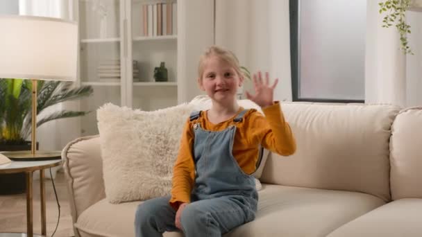 カメラを振っている小さな笑顔の白人少女の娘は オンライン会議ビデオコールバーチャルトークコンセプトで親戚との家庭のソファーコミュニケーションで歓迎挨拶を送ります — ストック動画