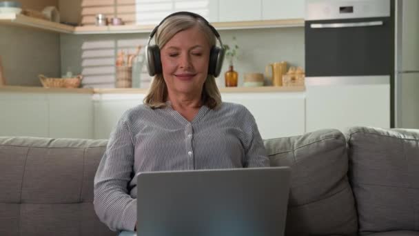 笑顔成熟した白人女性祖母のビジネスマンフリーランサーは ヘッドフォンで音楽を聞く自宅のラップトップにタイピングソーシャルネットワークコンセプト現代技術と高齢者 — ストック動画