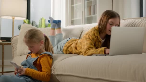 つの白人の子供の少女 姉妹の娘 リビングルームで異なるガジェットを使用して一緒に面白いビデオ子供や現代技術のコンセプトを見ているラップトップ携帯電話でオンラインゲームをプレイ — ストック動画