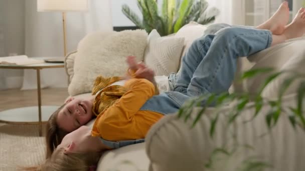 笑无忧无虑的白人女孩孩子们姐妹们的女儿们倒地躺在客厅沙发上玩耍玩耍放松玩耍嬉笑在家里过一个快乐的周末快乐的家庭 — 图库视频影像