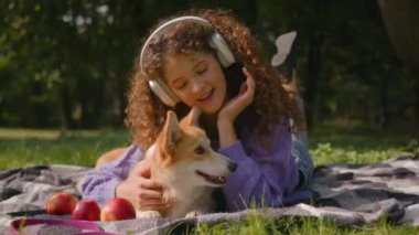 Mutlu gülümseyen beyaz 20 'li yaşlardaki genç kız öğrenci kulaklıkla müzik dinliyor. Parkta battaniyeye uzanmış, sevimli bir köpek yavrusu ve sahibinin sevgisini öpüyor.