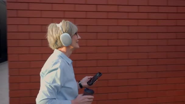 ケアフリーコーカサスシニア成熟した幸せなビジネスマンは 街の赤い壁の近くを歩くオフィスの朝のコーヒーを飲みに行く古いビジネス女性とヘッドフォンで音楽を聞きます — ストック動画