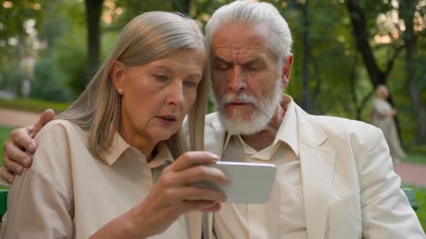 特写震惊忧心忡忡年事已高的养老金领取者男人白种人老两口坐在长椅外看手机屏幕在公园里读坏消息智能手机错误家庭不愉快的信息互联网 — 图库视频影像