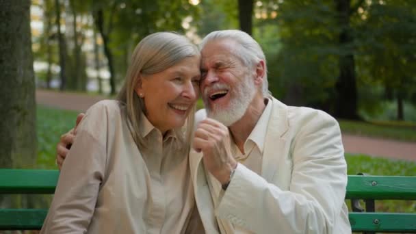 閉じた肖像画 歳の年金受給者の古いカップルがベンチに座って笑って 外の街公園を抱擁するカドルを話す 休憩週末一緒にリラックスする ケアフリー年金老人のおばあちゃん — ストック動画