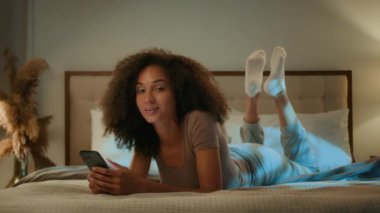 Mutlu Afro-Amerikalı genç kadın 20 'li yaşlarda kıvırcık saçlı güzel kadın gece yatak odasında rahat bir şekilde uzanmış cep telefonuyla kameraya bakıp gülümsüyor.