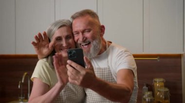 Olgun, emekli, beyaz bir çift, mutlu bir aile, mutlu bir büyükanne ve büyükbaba, internet üzerinden konuşuyor. Ev mutfağındaki cep telefonuyla, gülümseyen yaşlı bir kadın, yaşlı bir eş ve akıllı telefon kamerasına el sallıyor.