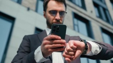 Beyaz tenli erkek işadamı ofisin dışında cep telefonu uygulaması kullanıyor düşünceli erkek işveren iş adamı, el kol saati kontrol zamanı, şehirde buluşma zamanı.