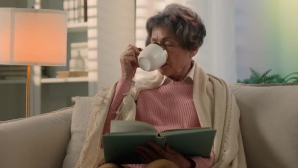 カップリーディング本とソファーの飲み茶コーヒーの毛布で覆われた穏やかな深刻なコーカサス古い女性高齢女性退職年金の祖母は家庭のソファーで小説の飲み物を読みます — ストック動画