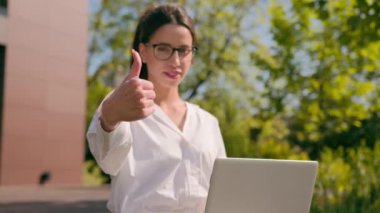Gülümseyen Kafkasyalı kadın iş kadını serbest çalışan bir öğrenci şehirde dizüstü bilgisayar kullanıyor. Başparmaklarını göstererek jest onayı veriyor. Reklam servisinin bilgisayar uygulamasını onaylamasını öneriyor.