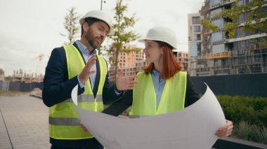 Caucasian two people colleagues coworkers builders architects engineers woman and man in helmets hardhats and vests discuss blueprint paper document plan work city outdoors urban building architecture clipart