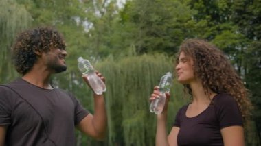 Fitness çok ırklı erkek kadın su içen dengeli vücut bakımı parkta sağlıklı enerjiyi gevşetiyor Arap erkek beyaz kız çift şehirde spor antrenmanından sonra dinleniyor