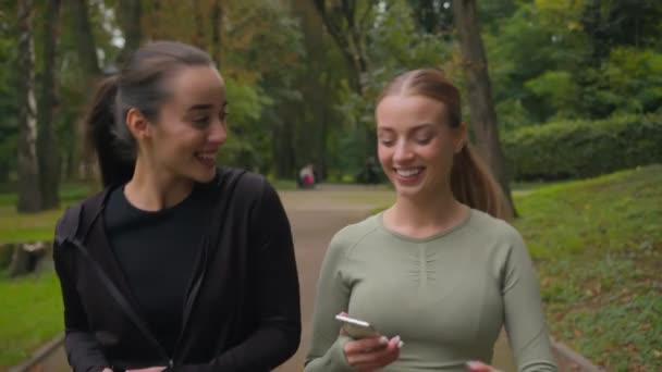 两个快乐的运动白人欧洲女人两个运动女孩在公园外面拿着电话散步 笑着积极适应运动女人跑步者与人交谈给予5个很好的团队精神女人友谊 — 图库视频影像